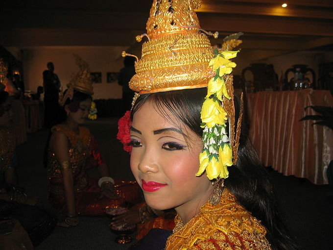 This is a current-day photo of a female Khmer dancer in Siem Reap in profile view. She wears a yellow hat and dramatic eye and lip makeup.