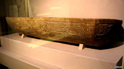 Photograph depicts an Uruk trough with carvings inside of a museum display case.