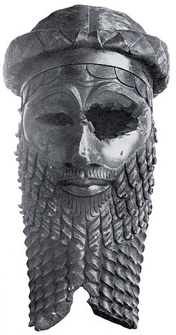 Photo portrays a bronze rendering of the face of an Akkadian ruler with strong features and a disfigured eye. The figure has an imposing beard and wears a headband.