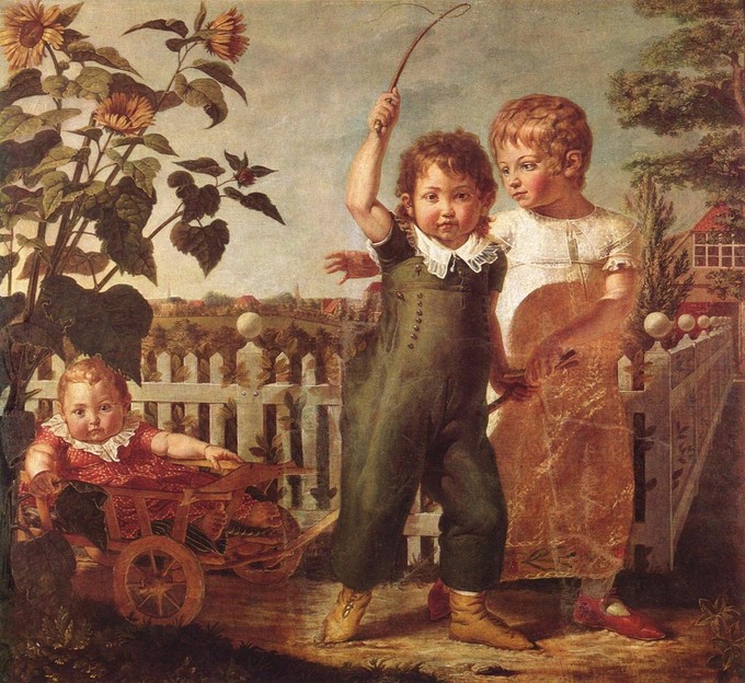 Two children are pulling a baby in a wagon next to a white picket fence. The baby and one of the children stares at the viewer. The other child looks back at the baby.