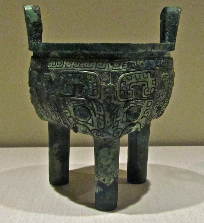 Photograph of a cooking pot that stands upright on three legs. It is covered with intricate carvings.