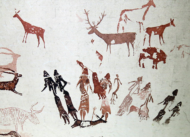 Prehistoric rock art depicts human figures surrounding an animal that has been speared. Other large game with horns and antlers surround the human figures.