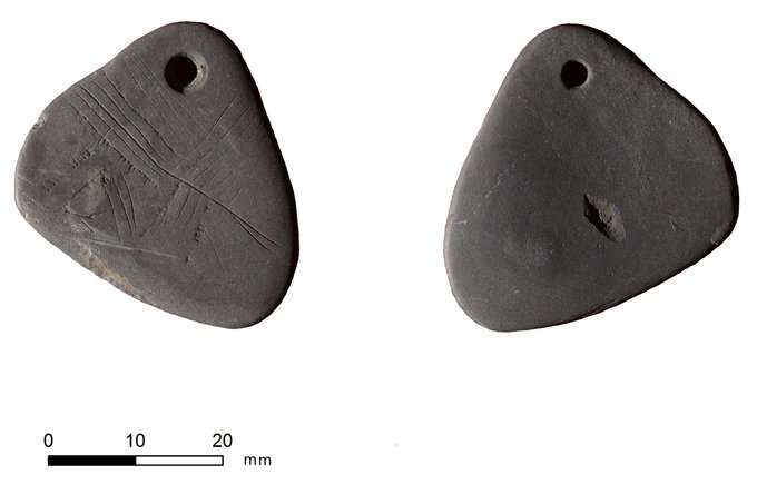 Photo depicts front and back view of a shale pendant. The pendant is shaped like a triangle with rounded edges. There are a series of lines etched into the stone.