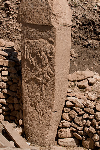 Photo shows mud brick t-shaped pillar with animals etched into it. It rests against the remains of a stone wall.