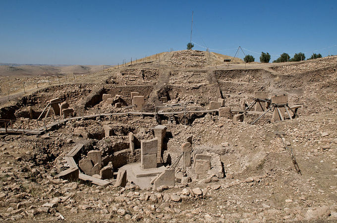 Photograph depicts ruins of Göbekli Tepe. The photo features a mound of dirt and rock with the remains of pillars and slabs that once formed a sacred structure.