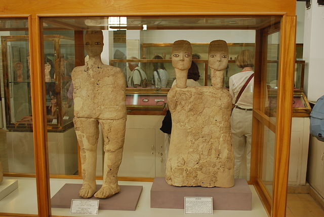 Photograph depicts statues and busts inside a display case at the Jordan Museum. On the left, is a full-length statue made from read and limestone. On the right is a two-headed bust made from the same materials.