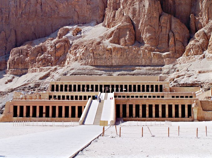 Photo depicts the Hatshepsut Temple, a structure of three layered terraces reaching 97 feet tall. Each story is articulated by a double colonnade of square piers. Behind the temple are the tall cliffs at Deir el Bahari.