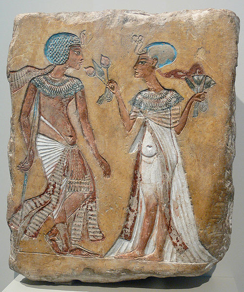 Painted relief depicts a man and a woman walking in a garden. Each wears a blue cap and is dressed in white, the man wears a long skirt, the woman wears a dress. Both wear jewelry on their necks. The woman holds flowers. The man holds a staff or walking stick.
