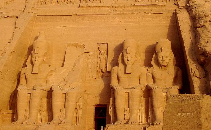 Four repeating statues of Ramse. The second statue has crumbled. The bearded pharaoh sits on a throne, hands on his thighs wearing a headdress with a cobra.