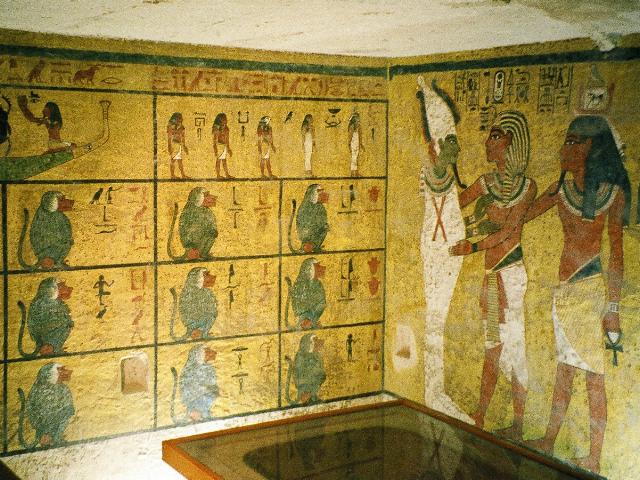 Photograph of Tutankhamun's burial chamber walls. Photo features the portion of the north wall decorated with a mural of King Tut embracing Osiris, clad in a white robe and hat. The western wall is decorated with illustrations that represent Egyptian beliefs about the afterlife, including twelve baboons, a boat, a beetle, and an assortment of gods.