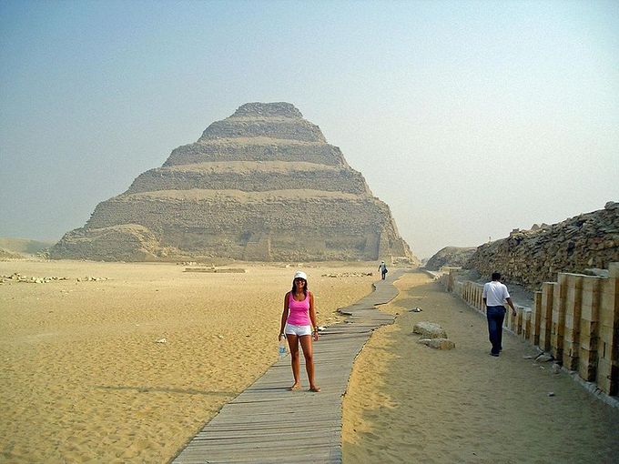 Photo depicts a tiered mud-brick pyramid in background with a woman posing in the foreground.