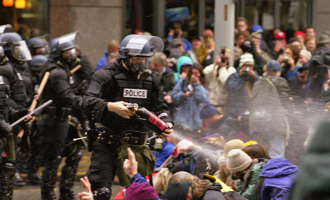 An armed policeman sprays tear gas into a group of protestors.