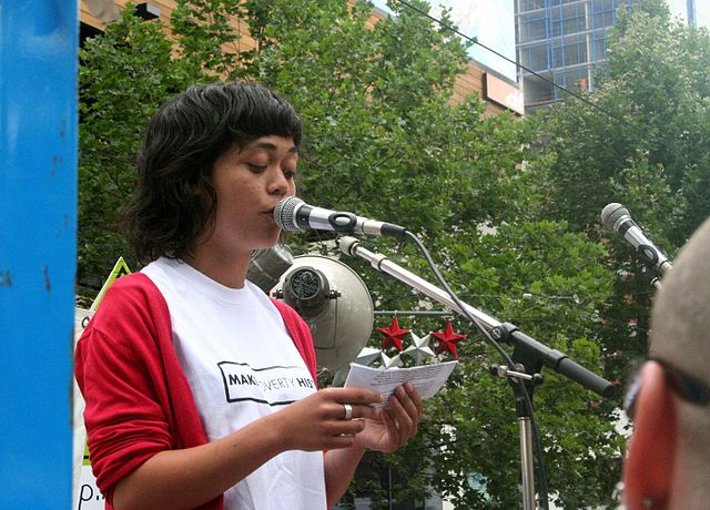 A picture of a young woman talking into a microphone during a public speech at a park.