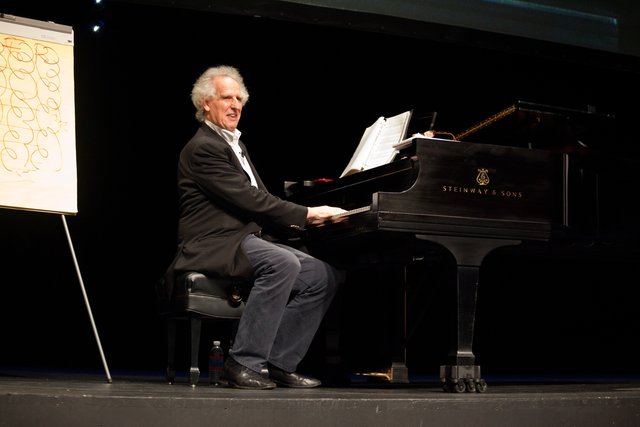 Benjamin Zander plays a piano on stage and connects with the audience.