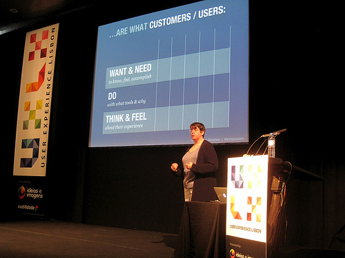 A woman stands on a stage and discusses a PowerPoint slide that breaks down customers wants and needs.