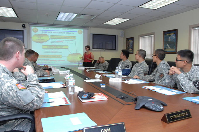 A group of soliders sits around a table while a woman discusses a PowerPoint presentation at the front of the room.