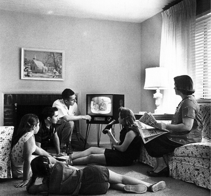 A family of six is gathered around a television set. The mother watches from the couch, while the father, the two sons, and the two daughters sit on the floor near the TV.