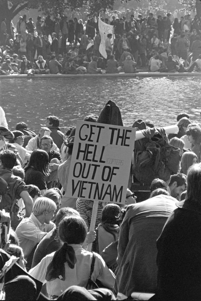 The photograph shows a large crowd of protesters. One holds a sign that reads 