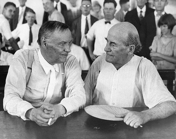 Photograph of Clarence Darrow and William Jennings Bryan sitting next to each other and conversing with each other in a courtroom. Clarence Darrow smokes a cigarette and William Jennings Bryan holds a fan.
