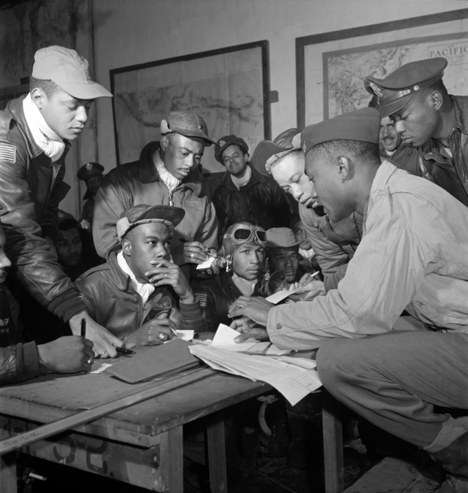 The photograph shows several Tuskegee airmen huddled around a table, discussing military plans.