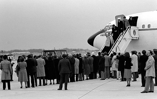 The photograph shows former hostages walking down a flight of steps to exit an official plane; a crowd of people waits for them on the ground.