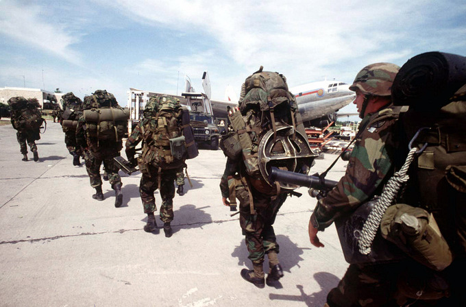 Seven armed soldiers carry their gear towards a plane.