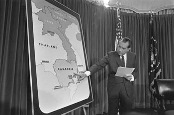 The photograph shows Nixon standing next to a large map of a portion of southeast Asia. He is pointing at Cambodia as he speaks.