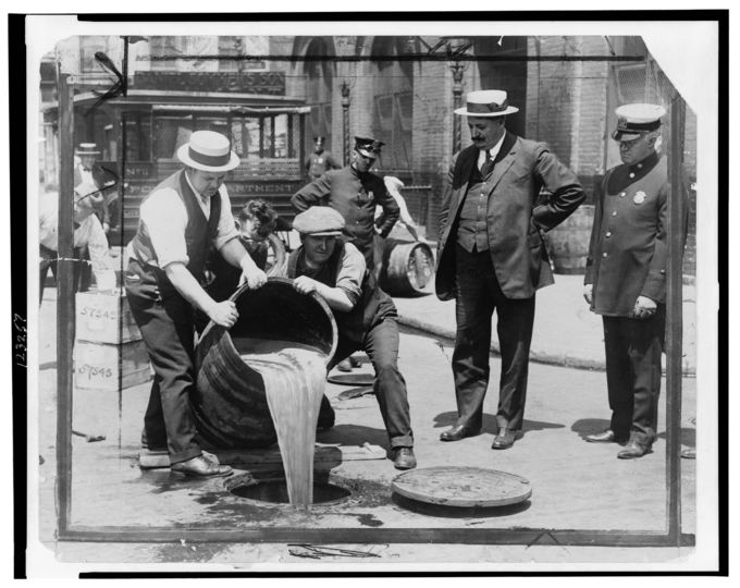 Photograph of three men dumping a barrel full of alcohol down a manhole as a number of men (including three officers) look on.