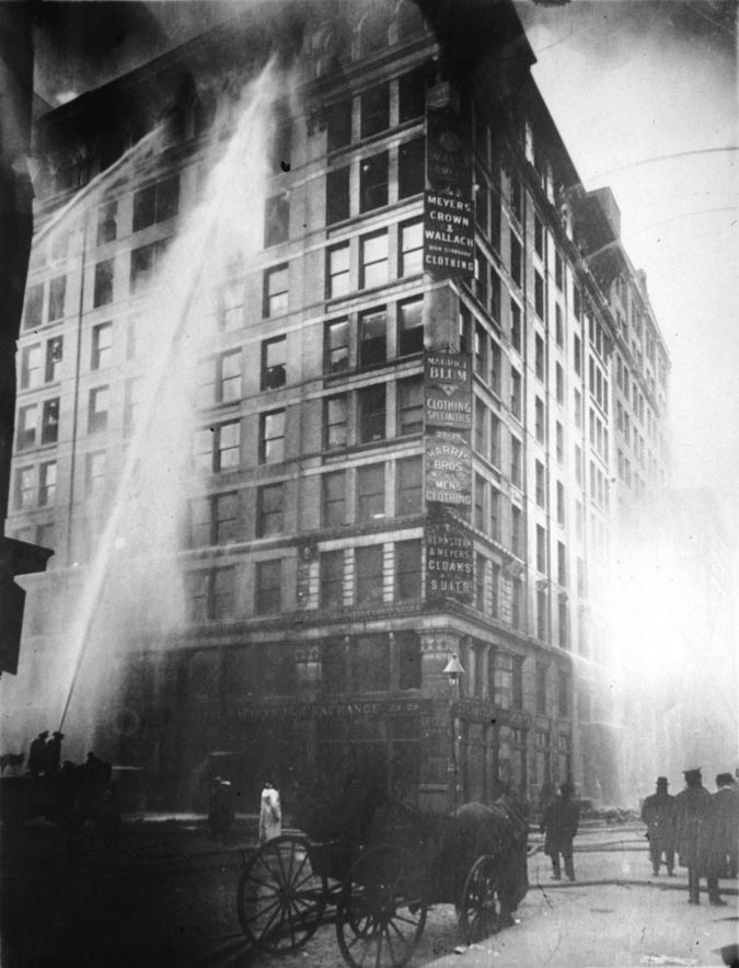 The photography shows firefighters attempting to put out the fire at the Triangle Shirtwaist Factory. The building is covered in ash.
