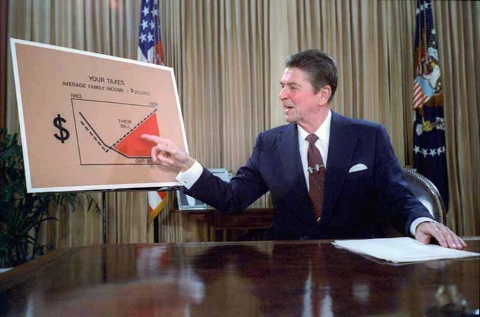 Ronald Reagan sits a desk. As he speaks, he points to a poster about tax reduction.