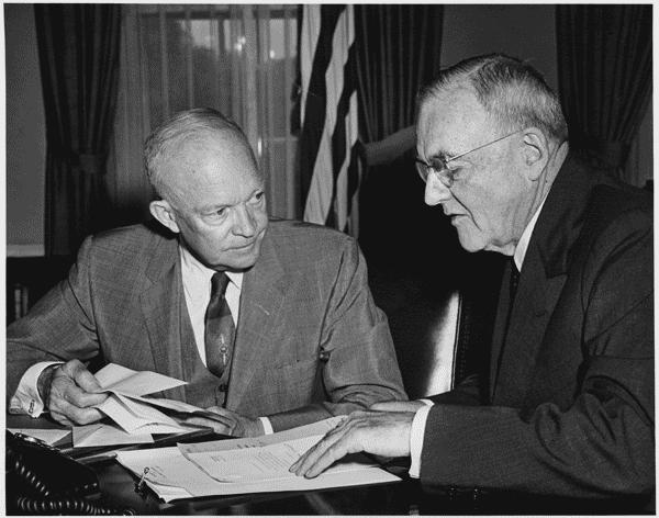 A photo of Secretary of State John Foster Dulles, right, shown here with President Eisenhower in 1956, sitting at a desk looking over papers.