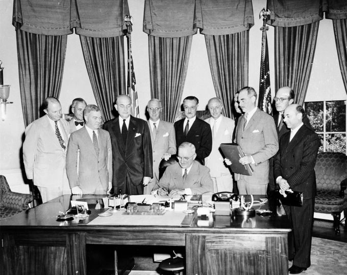 Photo of U.S. President Harry S. Truman signing the North Atlantic Treaty seated at the desk in the Oval Office with eleven men standing behind him.