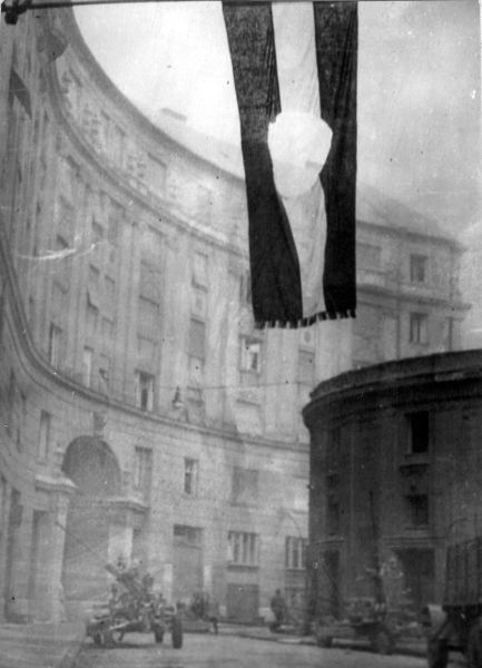 A photo of a Soviet Union flag with the communist coat of arms cut out hanging over a street. Military vehicles can be seen in the background.