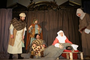 six actors on a stage. Central is a kneeling woman holding a man who appears passed out or dead. Around her kneel and stand four other men, dressed in medieval garb.