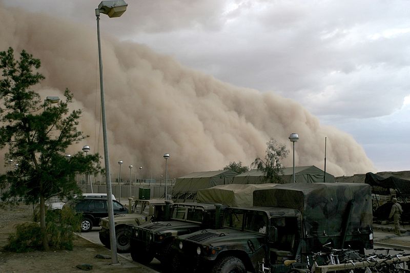 A large sandstorm blowing in over a parking lot in Iraq. The sand cloud is three or four times the height of the army trucks parked in the lot.