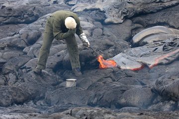 Scientist in protective gear sampling lava using a rock hammer and a bucket of water. 