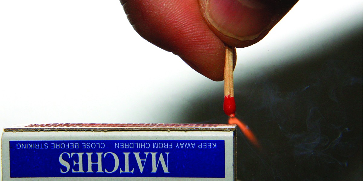 A match held in a person’s hand is ignited as it is scratched along the rough surface of a match box.