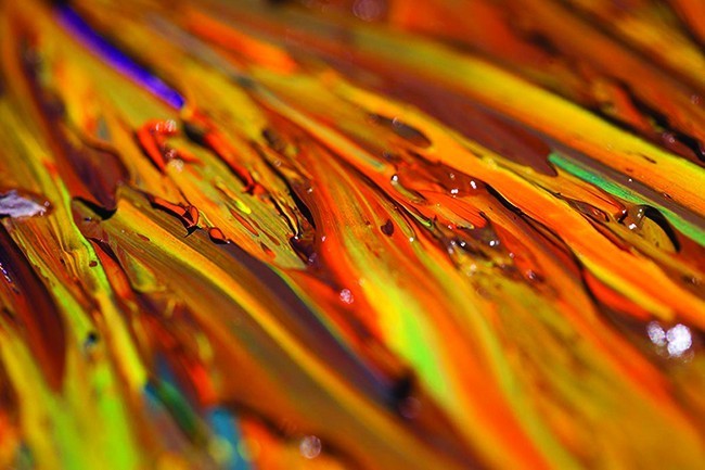 A photograph is shown of a portion of an oil painting which reveals colors of orange, brown, yellow, green, blue, and purple colors in its strokes. A few water droplets rest on the surface.