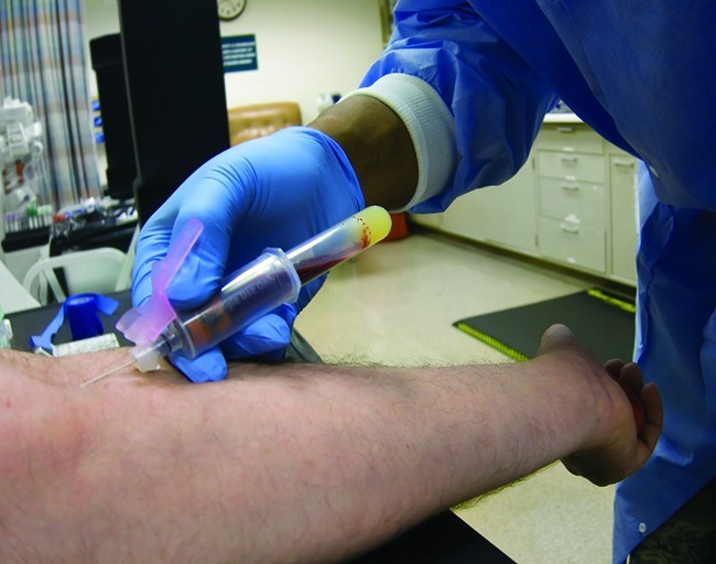 A photograph shows a person’s hand being held by a person wearing medical gloves. A thin glass tube is pressed against the persons finger and blood is moving up the tube.