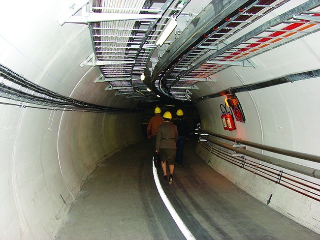 A tunnel of concrete with rails on the ground and tubes and wires running along the wall. Two people walk along the tunnel.