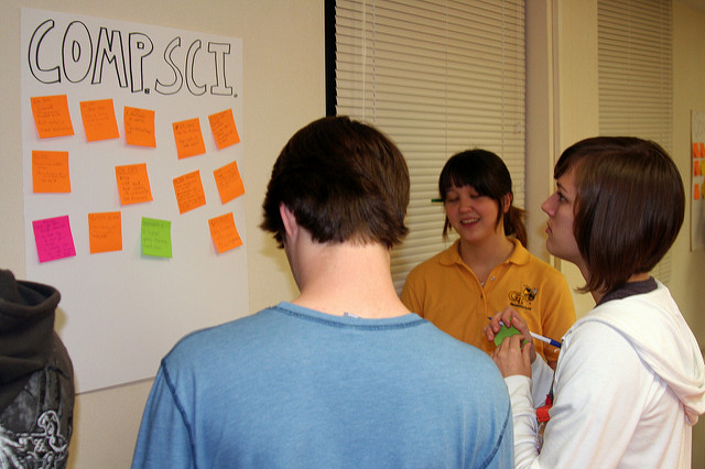 Photo of a group of students standing around a poster on the wall, where they're adding post-it notes with handwriting on them