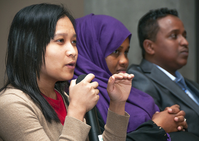 Photo of three people from ethnically diverse backgrounds sitting in a row. The woman closest to the camera holds a microphone, a woman in a burka sits next to her, and a man in a suit is at the end.