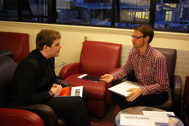 Two men sitting in leather chairs, leaning towards each other, both with papers on their laps. One is explaining something to the other, who is listening closely.