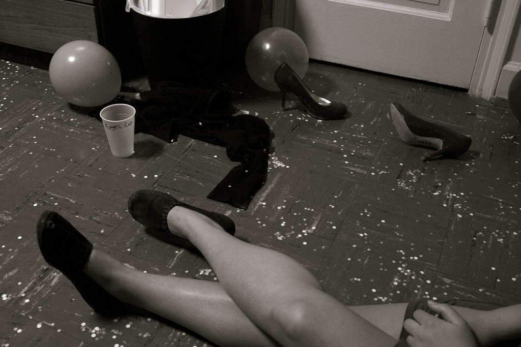 Photo of a woman's legs. Though mostly out of the frame, she appears to be sitting on the floor at a party. Next to her, on the floor, is a pair of high-heeled women's shoes, a party balloon, a plastic beer cup. 