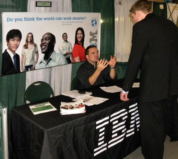 A student talking with a recruiter at an IBM booth. A sign behind the booth shows portraits of people from all ethnicities and the words Do you think the world can work smarter?