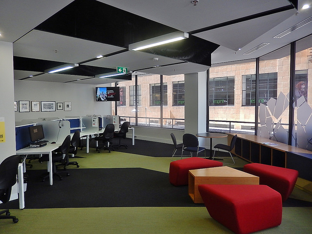 Photo of a modern workspace, with curved computer desks along one wall, movable red cushions and tables, and wide windows