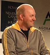 Photograph of a seated Marc Anderson, smiling away from the camera and wearing an adidas zip-up jacket.