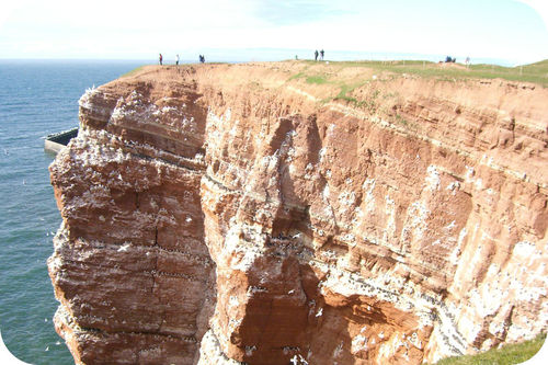 Several people standing on a massive cliff made of sandstone overlooking the ocean. The sandstone has clear striations.