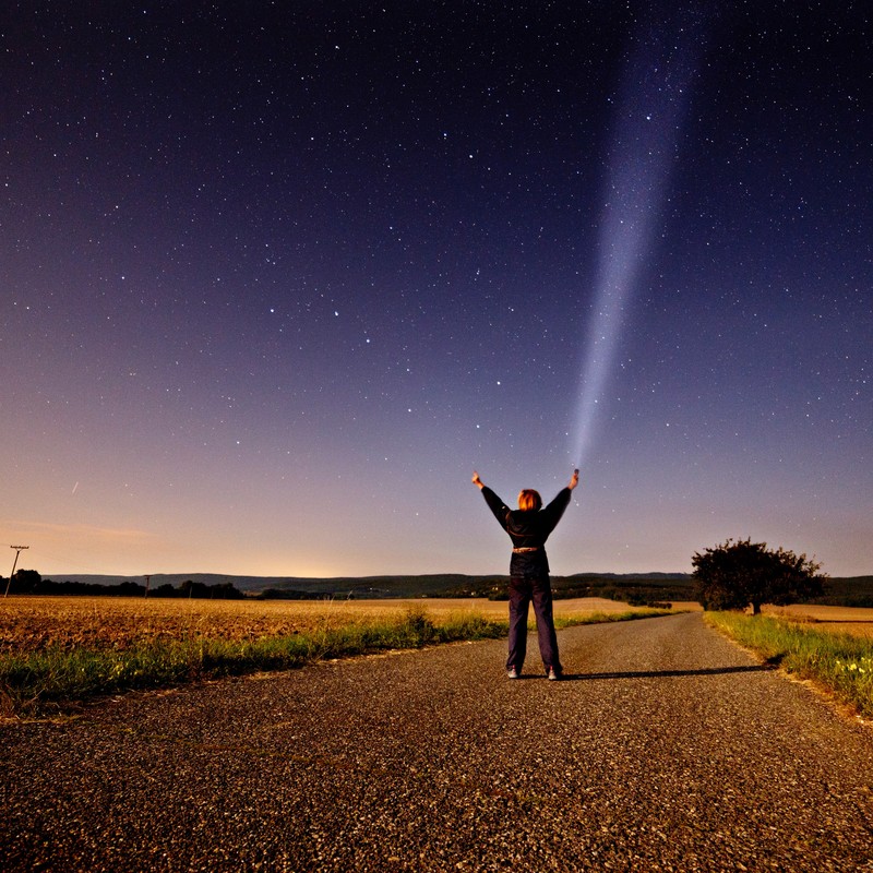 A woman stands in the middle of a country road at night and reaches toward the star-filled sky above. [Image: Maria Svecova, CC0 Public Domain, https://goo.gl/m25gce]