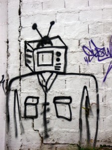 Photo of graffiti in black paint on a white wall, depicting a human figure with a television for a head
